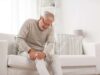 Älterer Mann sitzt mit Schmerzen im Knie auf dem Sofa und hält sich sein Knie.