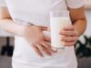 Eine junge Frau hält sich den Bauch vor Schmerzen und hält ein Glas Milch in ihrer Hand.