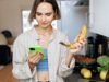 Junge Frau steht in der Küche und blickt auf ihr Smartphone. Neben ihr steht ein Laptop und Gemüse in einer Kiste.