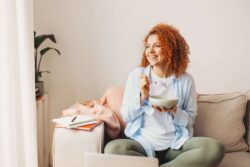 Junge Frau sitzt im Schneidersitz auf einem Sofa und isst etwas mit Gabel aus einer Schale.
