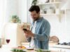 Ein junger Mann bereitet Essen in der Küche zu. Neben ihm steht ein Glas Rotwein. Er schaut auf sein Smartphone.