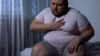 Junger Mann mit Übergewicht sitzt auf dem Bett und hält sich die Brust. Er hat Schmerzen im Brustkorb.