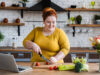 Eine junge, übergewichtige Frau bereitet in der Küche einen Salat zu. Vor Ihr auf der Arbeitsfläche steht ihr Laptop.
