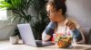 Eine junge Frau sitzt am Laptop, liest etwas auf dem Bildschirm und isst nebenbei einen Salat.