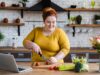 Eine junge, übergewichtige Frau bereitet in der Küche einen Salat zu. Vor Ihr auf der Arbeitsfläche steht ihr Laptop.